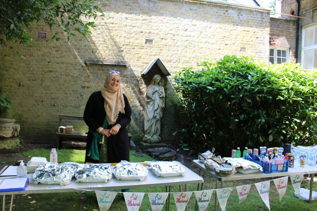 Muslim woman standing outside behind a table filled with food and drink and Eid banners.